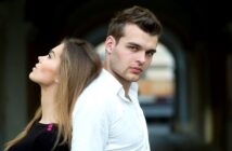 A man and woman standing back-to-back. The woman looks upward with her eyes closed, while the man looks forward with a neutral expression. They are in an outdoor setting with an archway in the background.