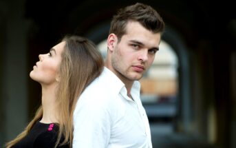 A man and woman standing back-to-back. The woman looks upward with her eyes closed, while the man looks forward with a neutral expression. They are in an outdoor setting with an archway in the background.