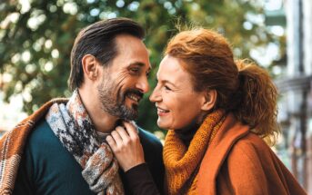 A happy couple dressed in cozy fall clothes smiles at each other while standing outdoors. The man wears a patterned scarf, and the woman wears an orange scarf and coat. Green foliage is blurred in the background.