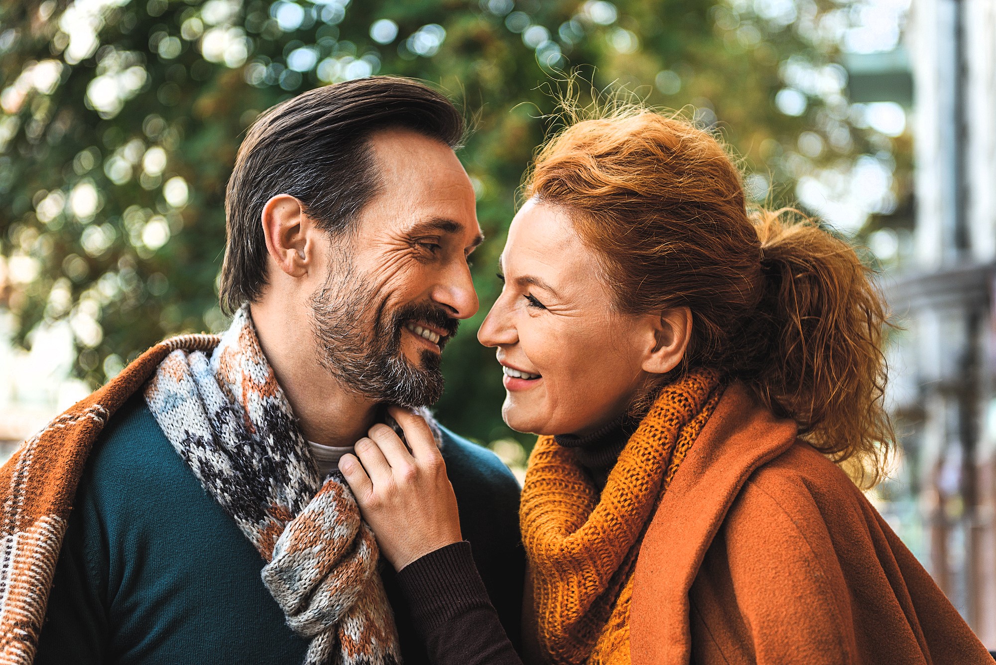 A happy couple dressed in cozy fall clothes smiles at each other while standing outdoors. The man wears a patterned scarf, and the woman wears an orange scarf and coat. Green foliage is blurred in the background.