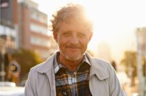 A man with curly hair and a beard smiles while standing outdoors in a city. He is wearing a plaid shirt and light jacket. The sun is setting behind him, creating a warm glow. Urban buildings are visible in the background.