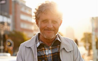 A man with curly hair and a beard smiles while standing outdoors in a city. He is wearing a plaid shirt and light jacket. The sun is setting behind him, creating a warm glow. Urban buildings are visible in the background.