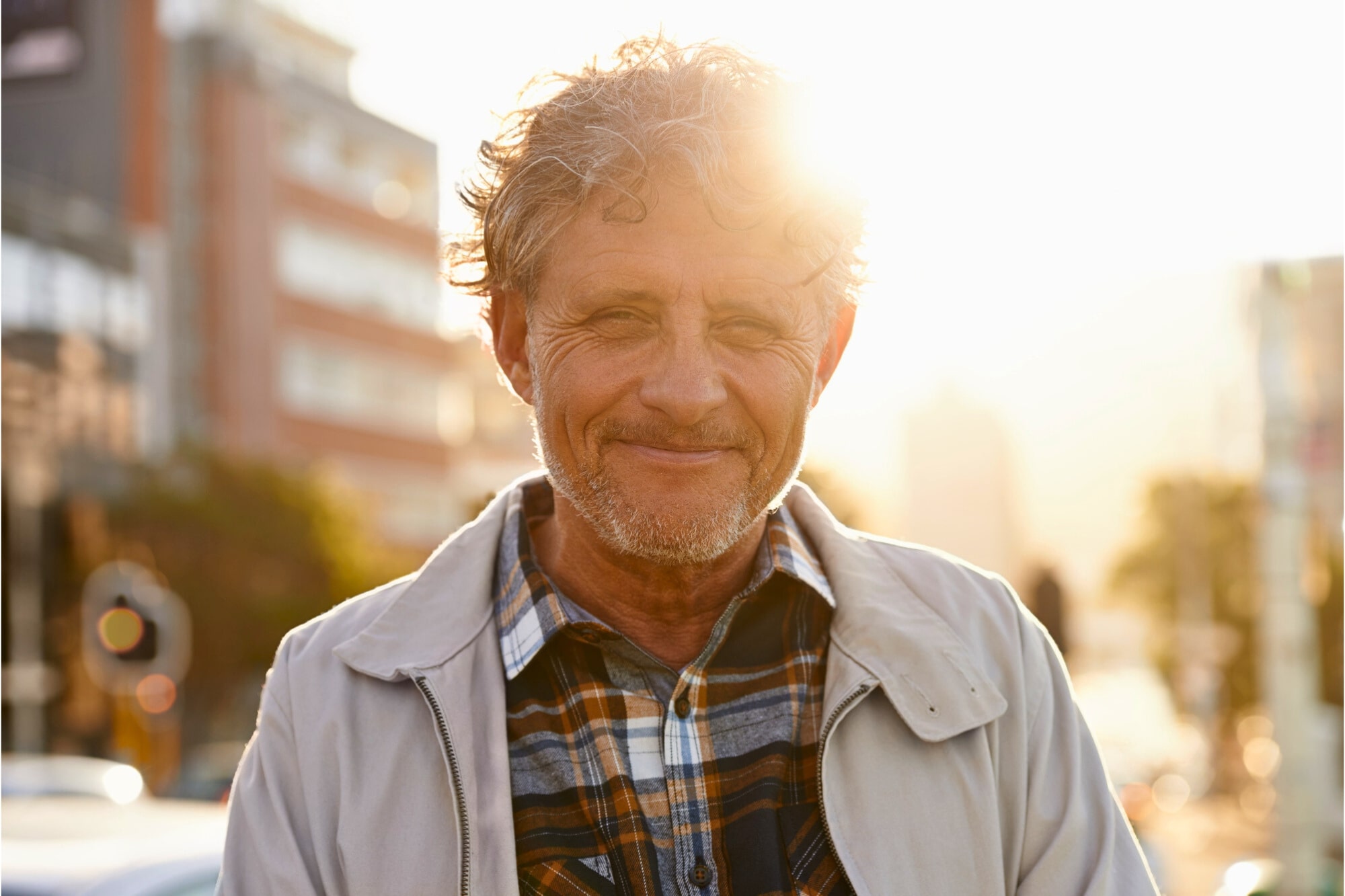 A man with curly hair and a beard smiles while standing outdoors in a city. He is wearing a plaid shirt and light jacket. The sun is setting behind him, creating a warm glow. Urban buildings are visible in the background.