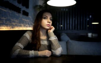 A woman with long hair sits at a dark wooden table in a dimly lit room, resting her chin on her hand. She gazes into the distance thoughtfully. The room has a modern, cozy ambiance with soft lighting.