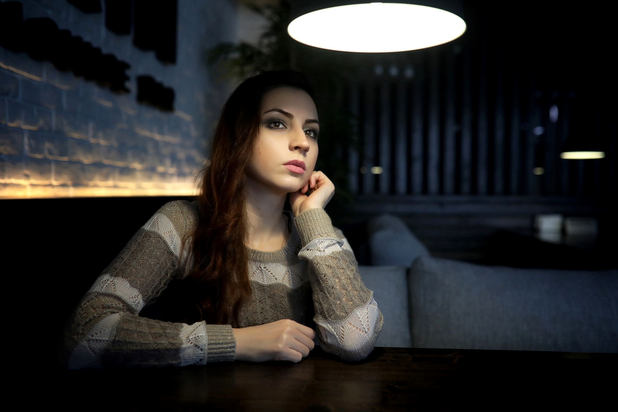 A woman with long hair sits at a dark wooden table in a dimly lit room, resting her chin on her hand. She gazes into the distance thoughtfully. The room has a modern, cozy ambiance with soft lighting.