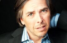 Man with gray hair and a light stubble, wearing a striped shirt and dark blazer, has a contemplative expression. He is looking slightly off-camera with a hand behind his head, set against a dark background.