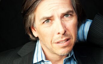 Man with gray hair and a light stubble, wearing a striped shirt and dark blazer, has a contemplative expression. He is looking slightly off-camera with a hand behind his head, set against a dark background.