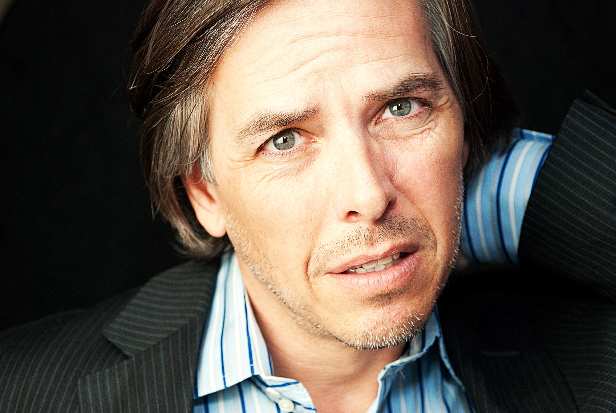 Man with gray hair and a light stubble, wearing a striped shirt and dark blazer, has a contemplative expression. He is looking slightly off-camera with a hand behind his head, set against a dark background.