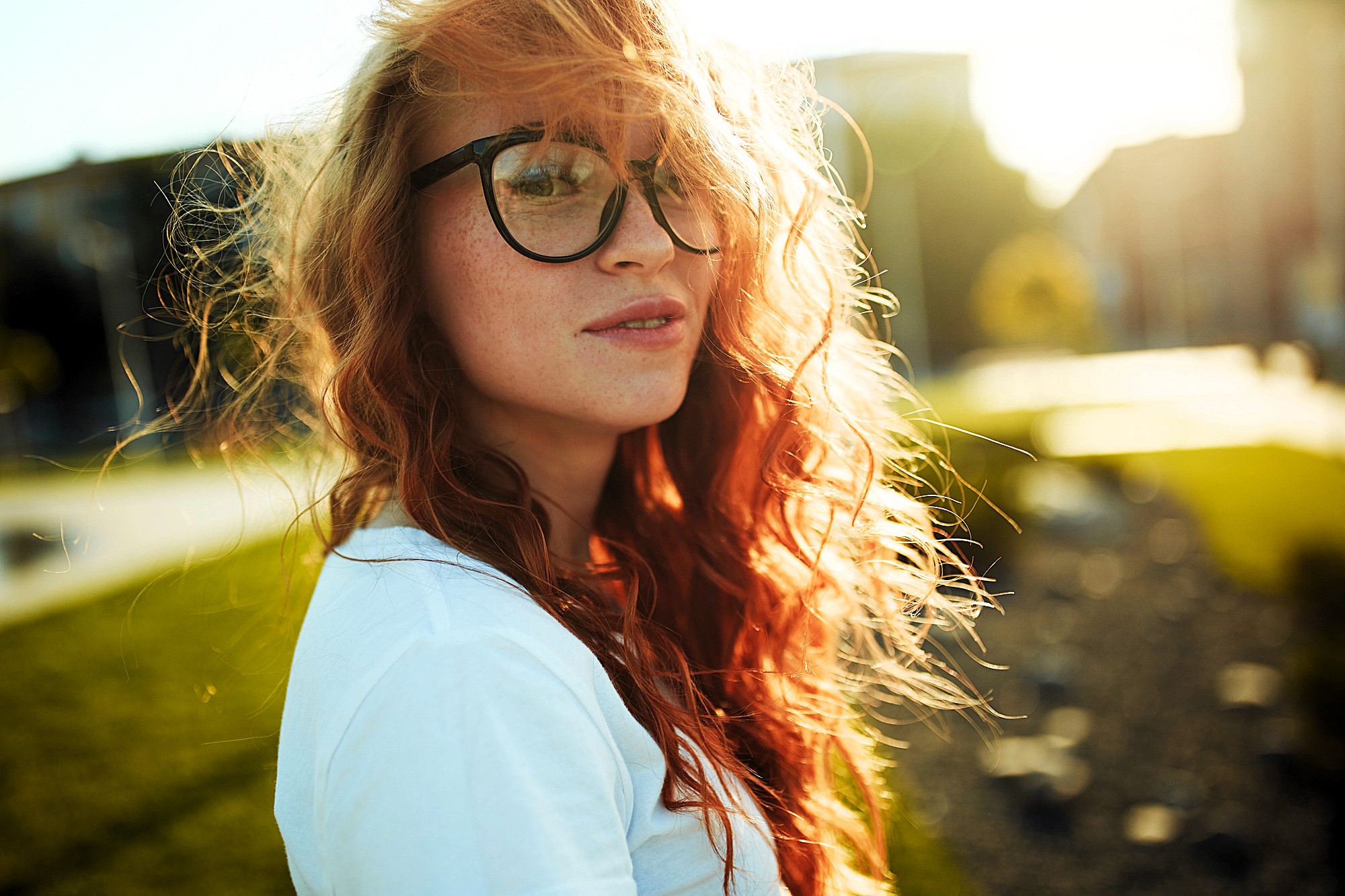 A young person with long, wavy red hair and black glasses stands outdoors. They are wearing a white shirt, and the sunlight creates a warm glow. The background features greenery and a blurred cityscape.