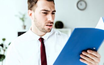 A man with a beard, wearing a white shirt and red tie, looks at a blue folder with a puzzled expression. Blurred plants and a clock are visible in the background.