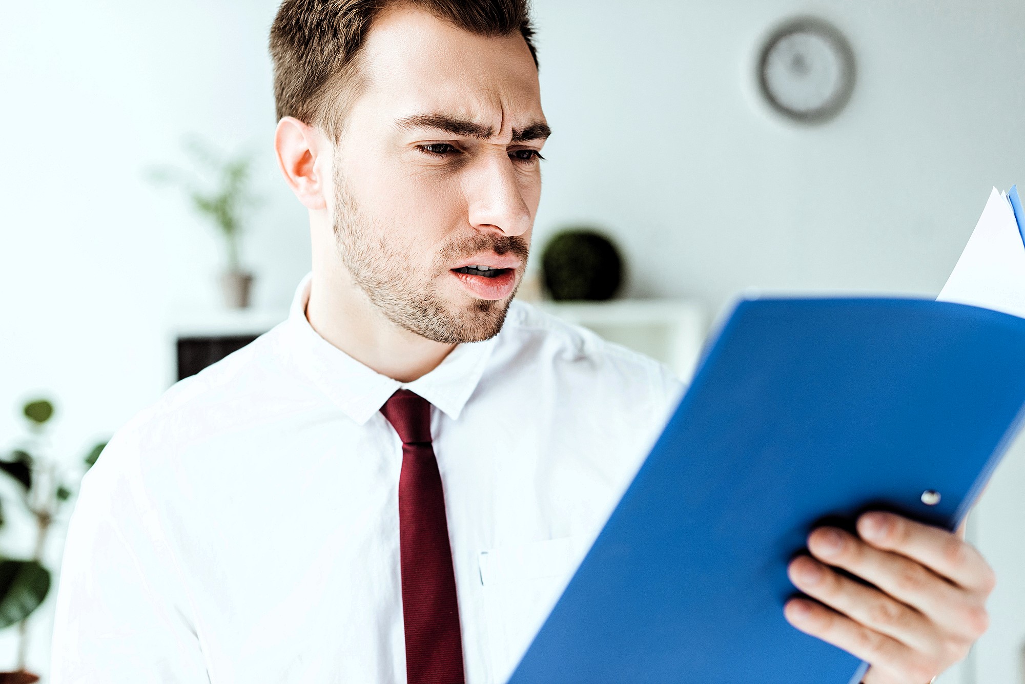 A man with a beard, wearing a white shirt and red tie, looks at a blue folder with a puzzled expression. Blurred plants and a clock are visible in the background.