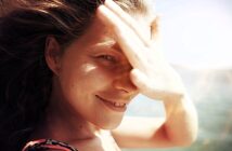 A woman smiles with her hand shielding her eyes from the sun. Her hair is blowing in the wind, and the background shows a clear blue sky and a glimpse of water. The sunlight creates a warm, natural glow on her face.