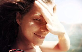 A woman smiles with her hand shielding her eyes from the sun. Her hair is blowing in the wind, and the background shows a clear blue sky and a glimpse of water. The sunlight creates a warm, natural glow on her face.