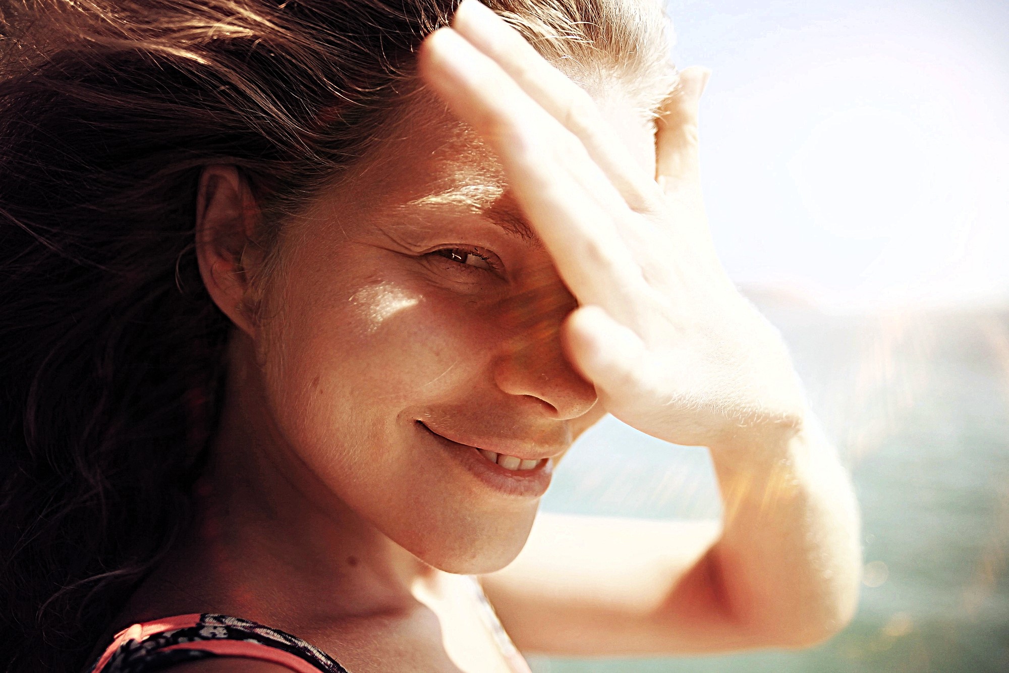 A woman smiles with her hand shielding her eyes from the sun. Her hair is blowing in the wind, and the background shows a clear blue sky and a glimpse of water. The sunlight creates a warm, natural glow on her face.