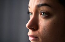 A close-up of a person's face in profile with tears streaming down. The lighting highlights their eyes and skin texture, conveying a sense of emotion.