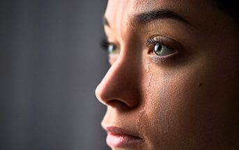 A close-up of a person's face in profile with tears streaming down. The lighting highlights their eyes and skin texture, conveying a sense of emotion.