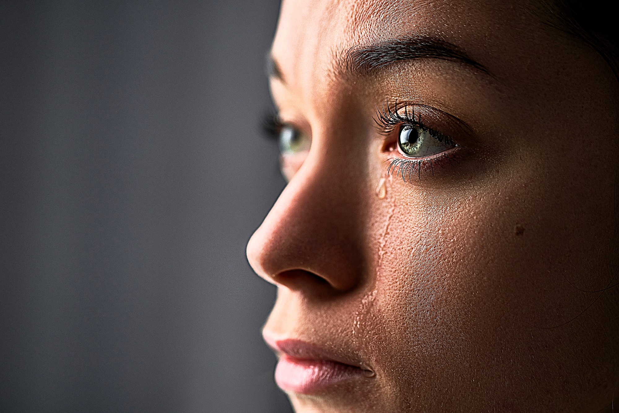 A close-up of a person's face in profile with tears streaming down. The lighting highlights their eyes and skin texture, conveying a sense of emotion.