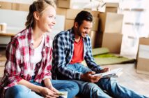 A smiling woman and man sit on the floor amidst moving boxes. Both are casually dressed in plaid shirts and jeans. The woman holds a phone, while the man holds a book. A coffee cup is visible in the background.