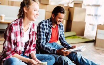 A smiling woman and man sit on the floor amidst moving boxes. Both are casually dressed in plaid shirts and jeans. The woman holds a phone, while the man holds a book. A coffee cup is visible in the background.