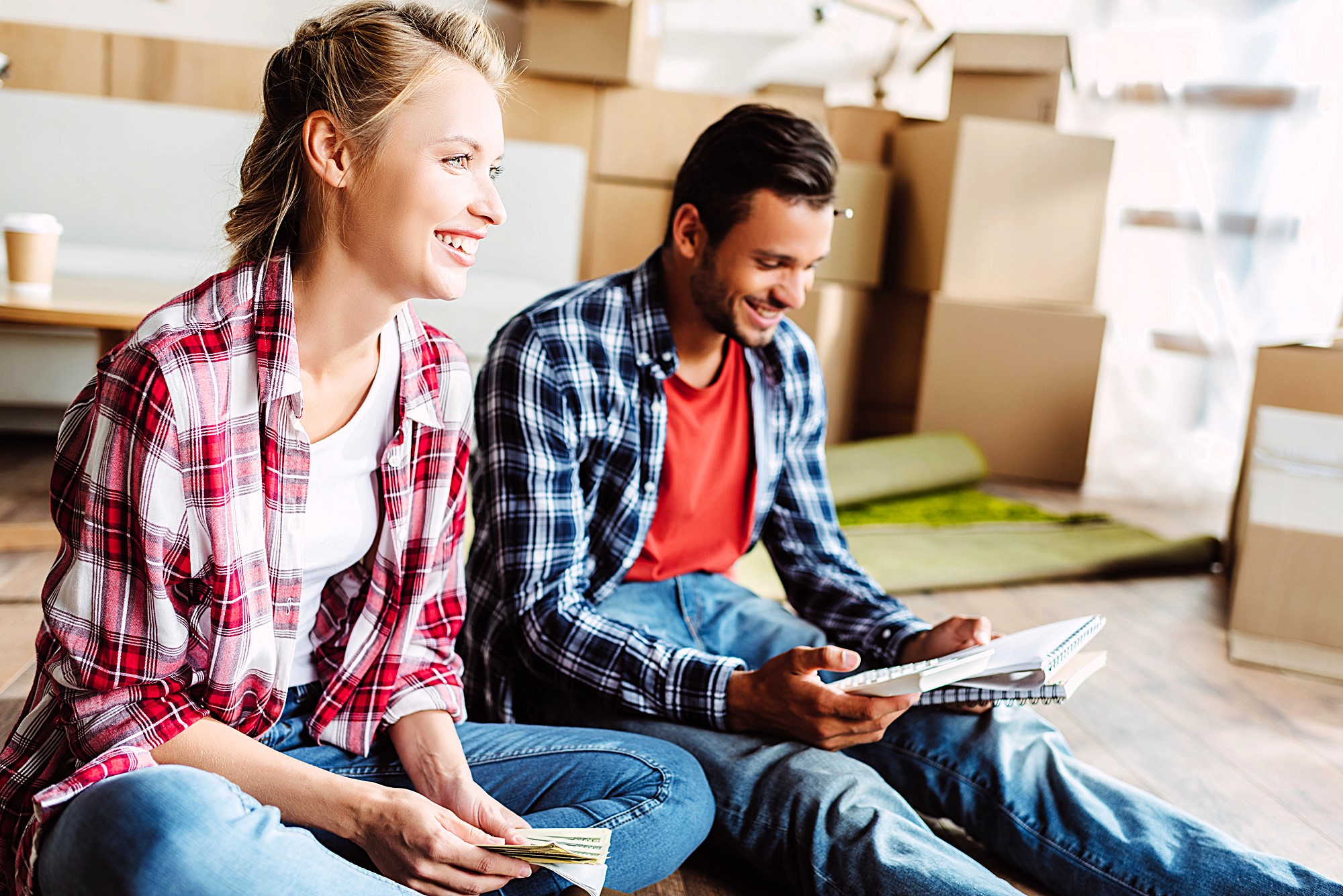 A smiling woman and man sit on the floor amidst moving boxes. Both are casually dressed in plaid shirts and jeans. The woman holds a phone, while the man holds a book. A coffee cup is visible in the background.