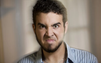 A man with curly hair and a beard appears angry, with his face scrunched up. He is wearing a striped shirt and is indoors, with a blurred background.