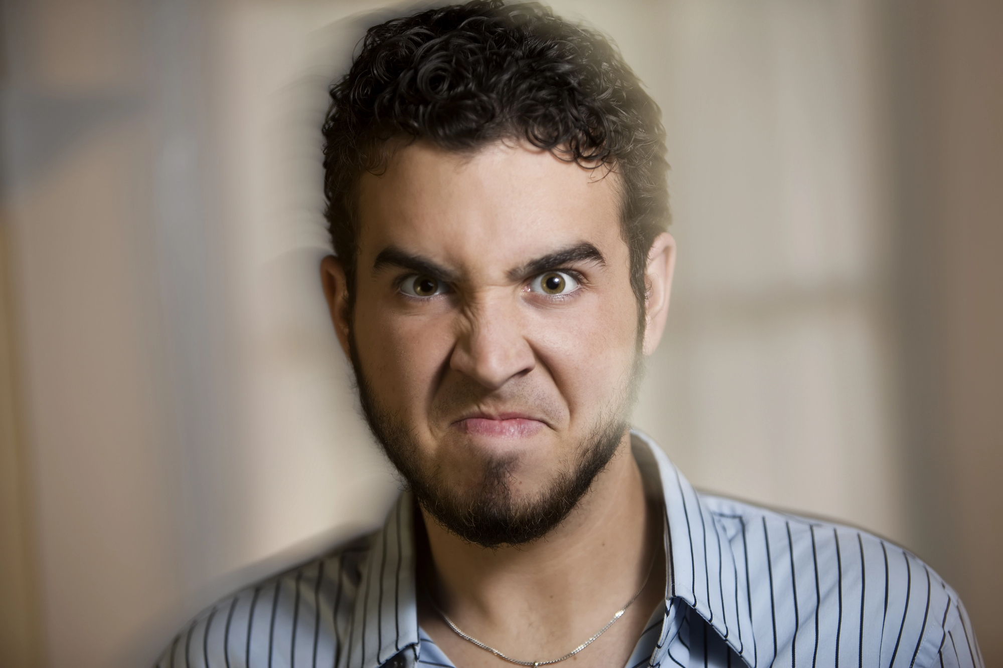 A man with curly hair and a beard appears angry, with his face scrunched up. He is wearing a striped shirt and is indoors, with a blurred background.