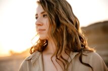 Woman with long, wavy brown hair gazes into the distance in a sunlit outdoor scene. She wears a beige shirt, and the warm sunlight highlights her face and hair, creating a serene and thoughtful atmosphere.