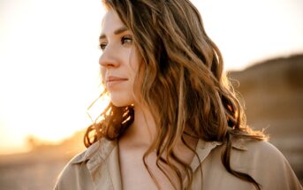 Woman with long, wavy brown hair gazes into the distance in a sunlit outdoor scene. She wears a beige shirt, and the warm sunlight highlights her face and hair, creating a serene and thoughtful atmosphere.