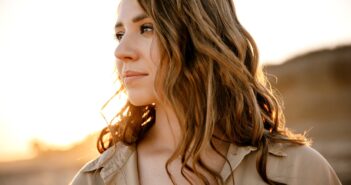 Woman with long, wavy brown hair gazes into the distance in a sunlit outdoor scene. She wears a beige shirt, and the warm sunlight highlights her face and hair, creating a serene and thoughtful atmosphere.