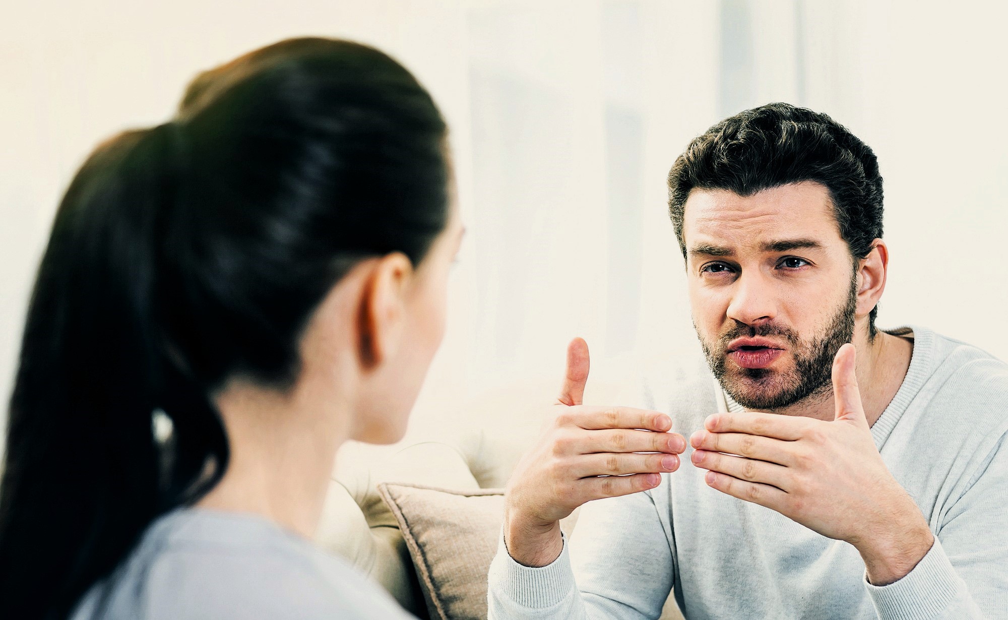 A man and a woman are engaged in a conversation indoors. The man is gesturing with his hands, appearing to explain or emphasize a point, while the woman listens attentively with her hair pulled back. They are seated on a light-colored couch.