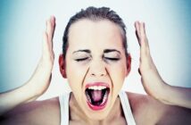 A woman with her hands near her head appears to be yelling, with eyes shut and mouth open wide. She has a neutral background, and her expression is intense, suggesting distress or frustration.