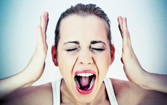 A woman with her hands near her head appears to be yelling, with eyes shut and mouth open wide. She has a neutral background, and her expression is intense, suggesting distress or frustration.