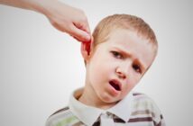 A young boy with short blonde hair and a striped polo shirt looks uncomfortable as a hand gently pulls his ear. The background is plain white.