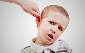 A young boy with short blonde hair and a striped polo shirt looks uncomfortable as a hand gently pulls his ear. The background is plain white.