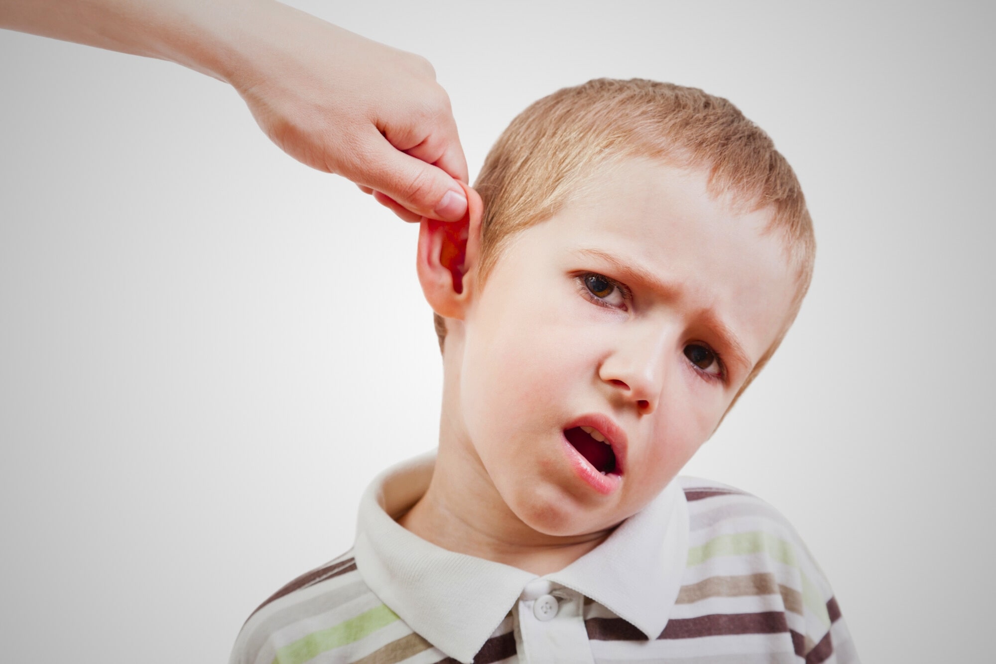 A young boy with short blonde hair and a striped polo shirt looks uncomfortable as a hand gently pulls his ear. The background is plain white.