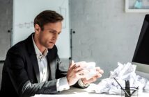 A man in a suit sits at a desk, looking thoughtfully at crumpled paper in his hands. More crumpled papers are scattered on the desk. A whiteboard with graphs is in the background, suggesting a work or office setting.