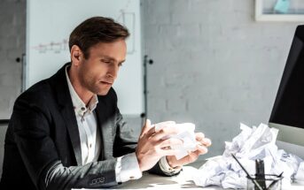 A man in a suit sits at a desk, looking thoughtfully at crumpled paper in his hands. More crumpled papers are scattered on the desk. A whiteboard with graphs is in the background, suggesting a work or office setting.