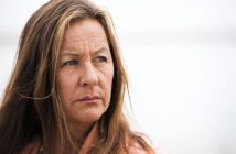 A woman with long brown hair and a serious expression gazes to the side against a blurred, light background. She is wearing a pink top and a beaded necklace.