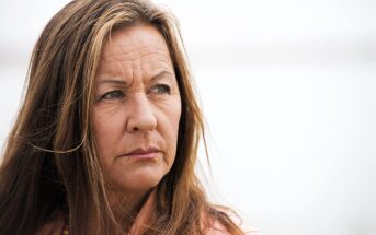 A woman with long brown hair and a serious expression gazes to the side against a blurred, light background. She is wearing a pink top and a beaded necklace.