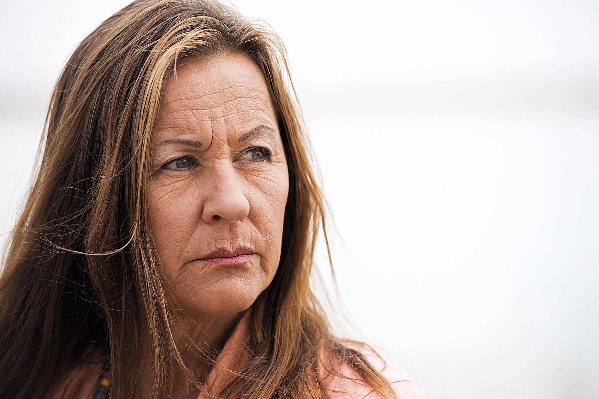 A woman with long brown hair and a serious expression gazes to the side against a blurred, light background. She is wearing a pink top and a beaded necklace.