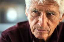 An older man with white hair and a serious expression looks to the side. He is wearing a dark red shirt. The background is blurred, suggesting an indoor setting.