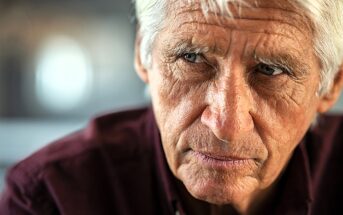 An older man with white hair and a serious expression looks to the side. He is wearing a dark red shirt. The background is blurred, suggesting an indoor setting.