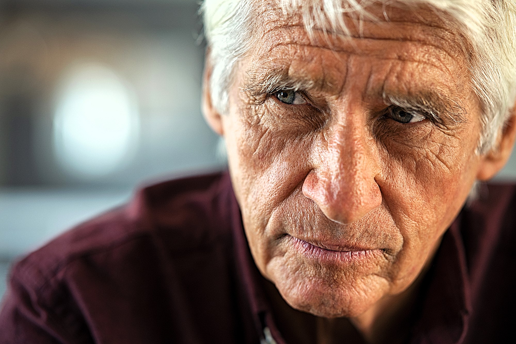 An older man with white hair and a serious expression looks to the side. He is wearing a dark red shirt. The background is blurred, suggesting an indoor setting.