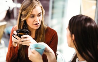 Two women sitting at a cafe table, holding mugs and engaged in conversation. One woman has long blonde hair and wears a red top; the other has dark brown hair and wears a light-colored top. They appear to be enjoying a casual chat in a sunlit setting.