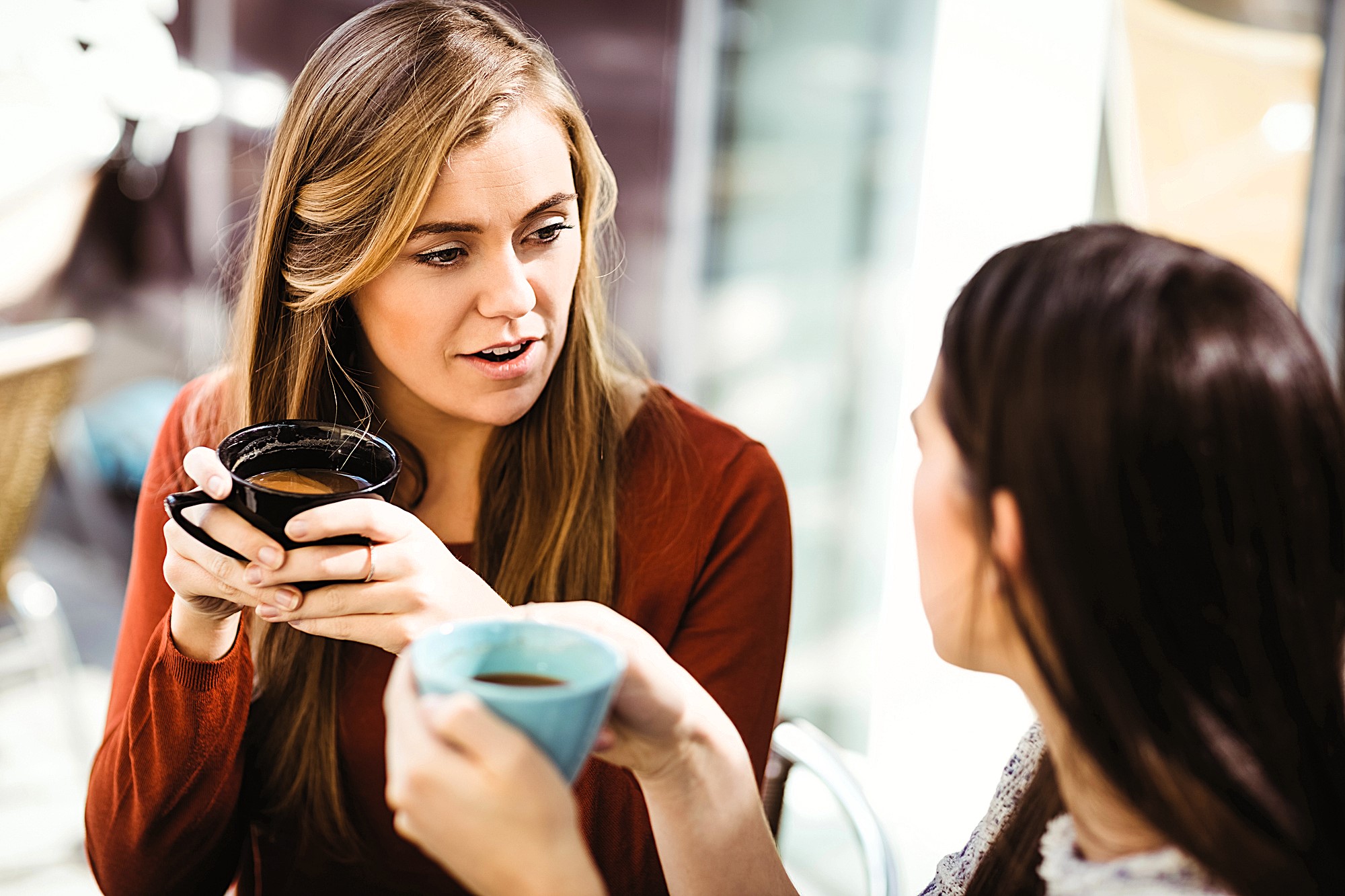 Two women sitting at a cafe table, holding mugs and engaged in conversation. One woman has long blonde hair and wears a red top; the other has dark brown hair and wears a light-colored top. They appear to be enjoying a casual chat in a sunlit setting.