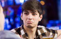 A young man with short dark hair, wearing a checkered shirt, sits at a table holding a glass of beer. He has a puzzled or unimpressed expression. The background is blurred, suggesting a bar or pub setting.