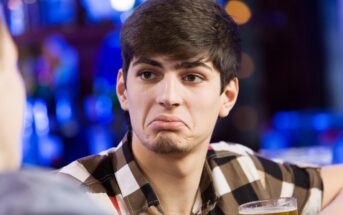 A young man with short dark hair, wearing a checkered shirt, sits at a table holding a glass of beer. He has a puzzled or unimpressed expression. The background is blurred, suggesting a bar or pub setting.