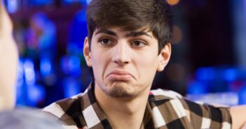 A young man with short dark hair, wearing a checkered shirt, sits at a table holding a glass of beer. He has a puzzled or unimpressed expression. The background is blurred, suggesting a bar or pub setting.