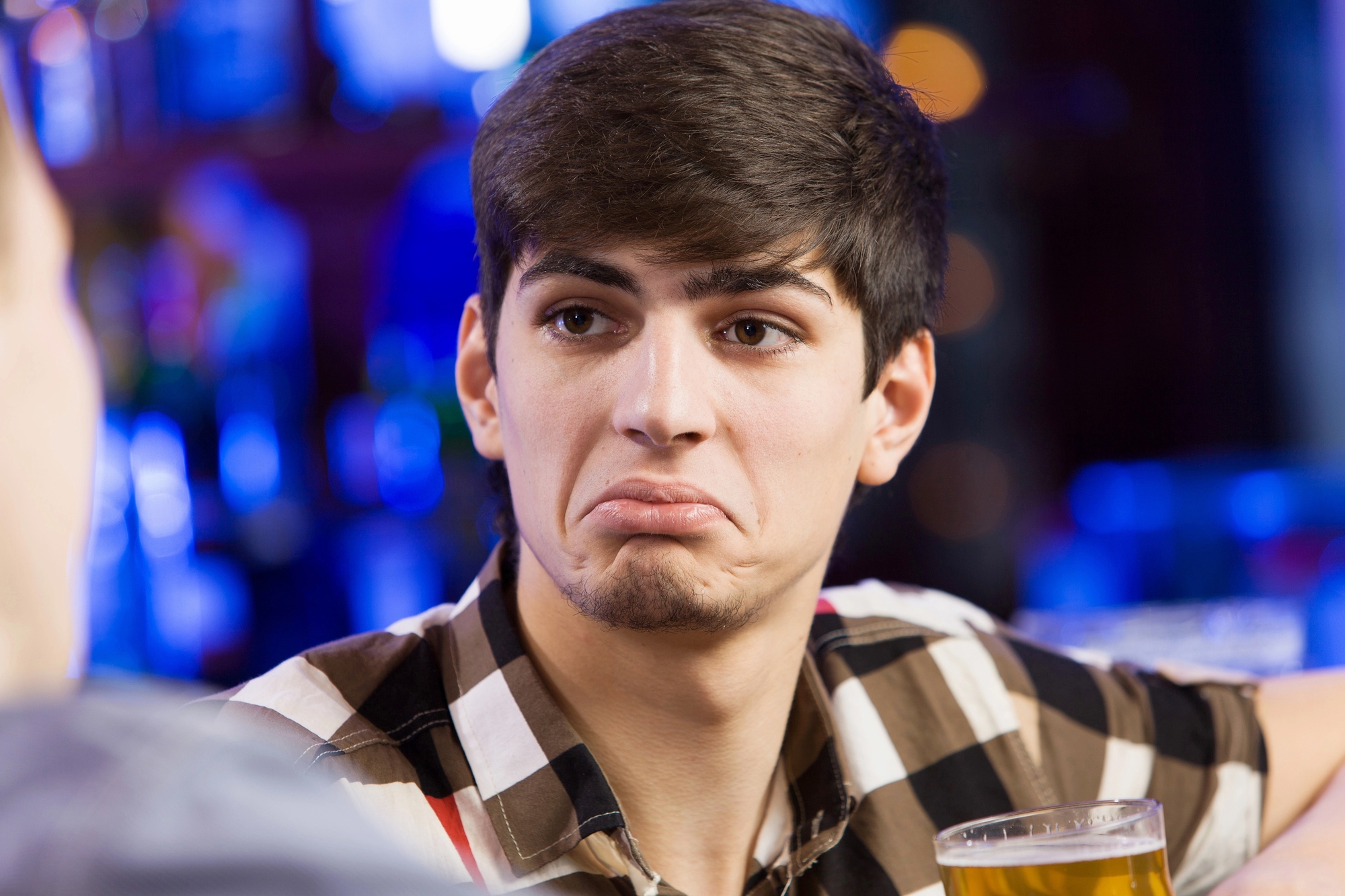 A young man with short dark hair, wearing a checkered shirt, sits at a table holding a glass of beer. He has a puzzled or unimpressed expression. The background is blurred, suggesting a bar or pub setting.