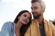 A woman with long brown hair leans her head affectionately on a man's shoulder. The man, with a beard and short brown hair, looks at her fondly. Both are outdoors in a sunlit setting with a clear sky in the background.